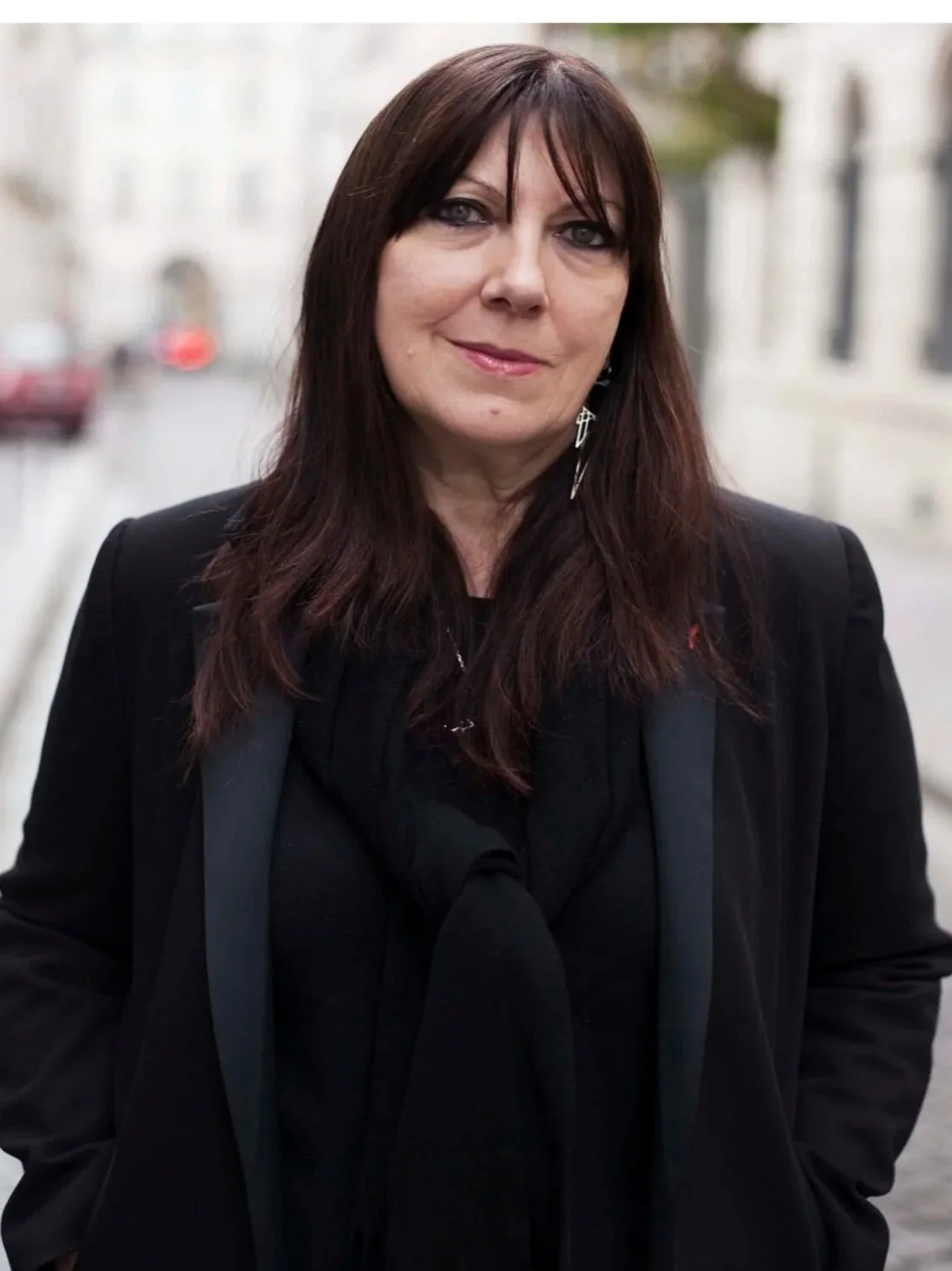 Une femme avec des cheveux bruns foncés et un maquillage sombre, portant un blazer noir et un foulard noir, se tenant dans une rue piétonne, probablement en ville.