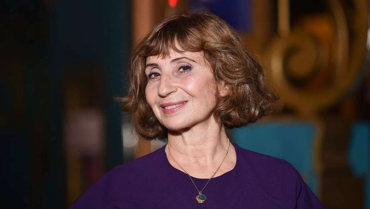 Une femme âgée avec des cheveux bruns courts, portant un chemisier violet et un collier doré avec un pendentif coloré, souriant légèrement.