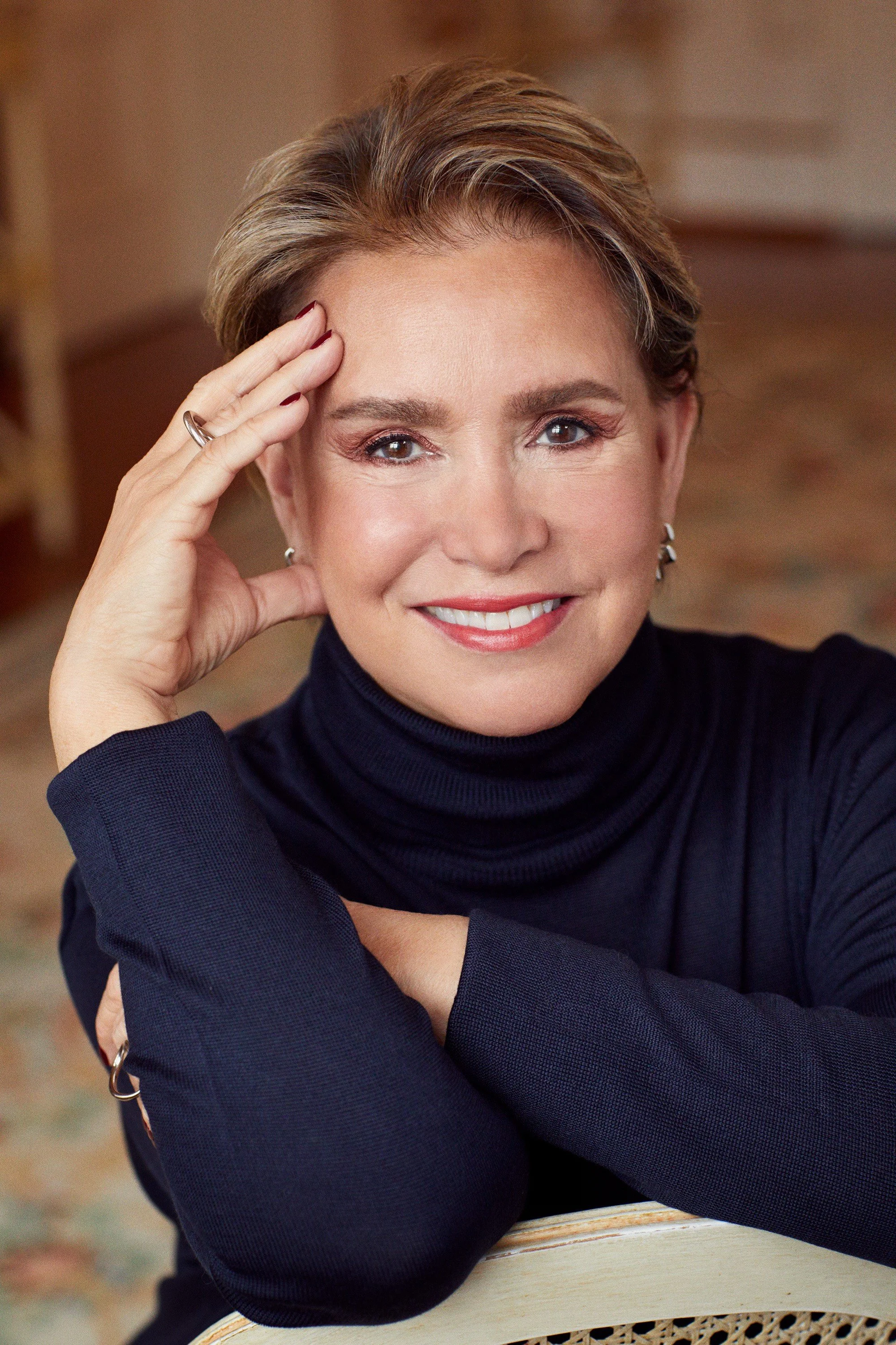 Portrait d'une femme souriante avec des cheveux courts, portant un haut à col roulé noir, posant avec la main sur la tête dans un intérieur chaleureux.