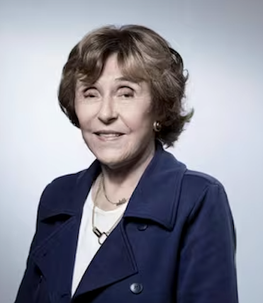 Une femme âgée avec des cheveux courts et bruns, portant une veste bleue, souriant en direction de l'appareil.