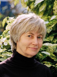 Une femme avec des cheveux courts et gris, portant un pull noir, posant devant des plantes vertes.