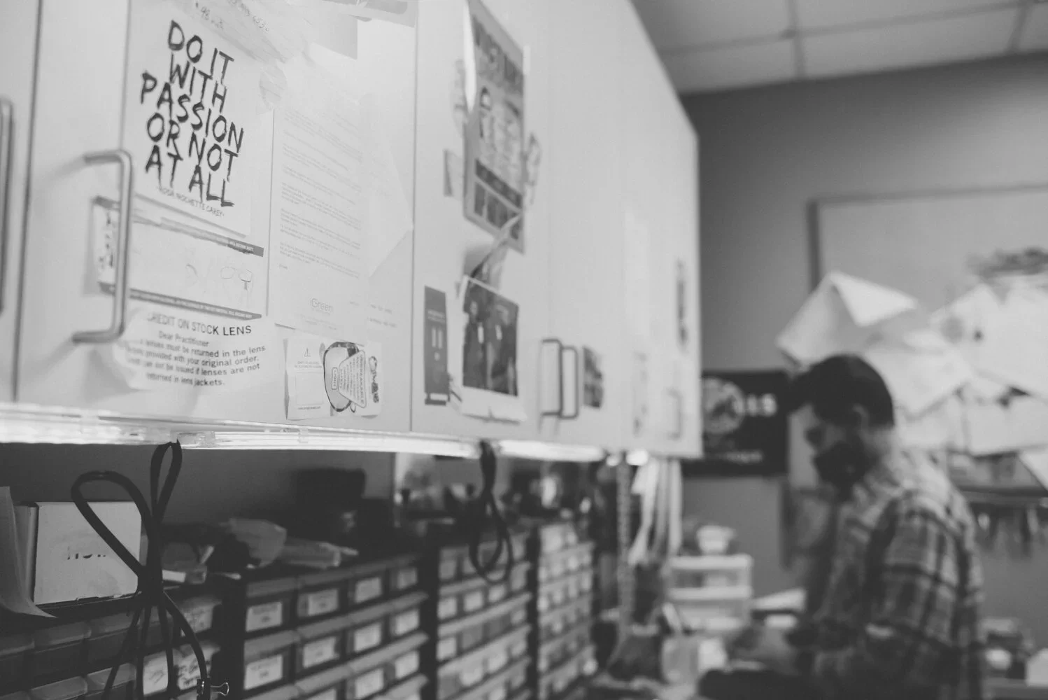 Black and white photo of person standing in a store aisle, wearing a face mask and goggles, surrounded by shelves with various items and signs.