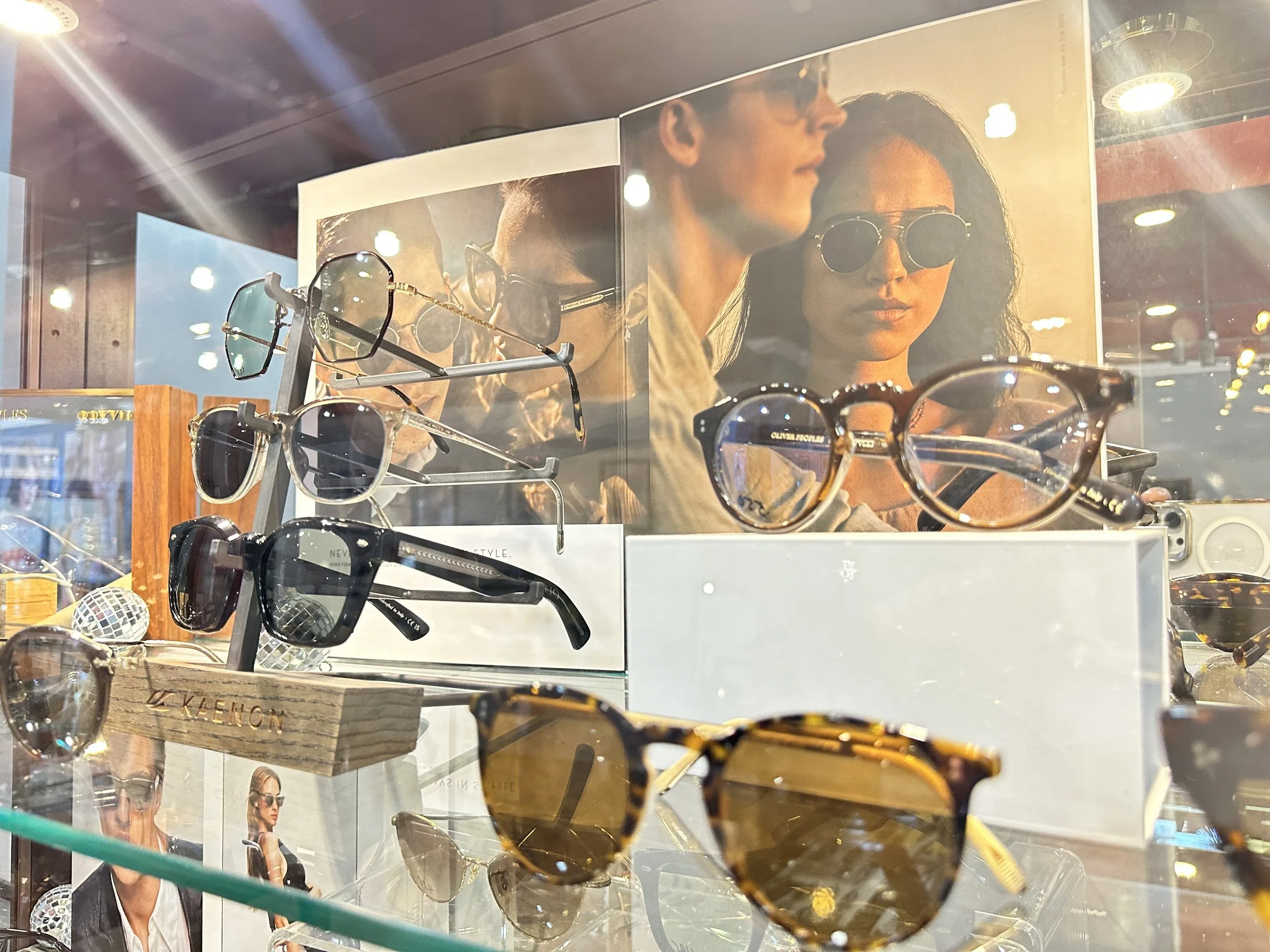 Display of various sunglasses including black, tortoiseshell, and metal frame styles inside a store window, with a large poster of a man and woman wearing sunglasses in the background.