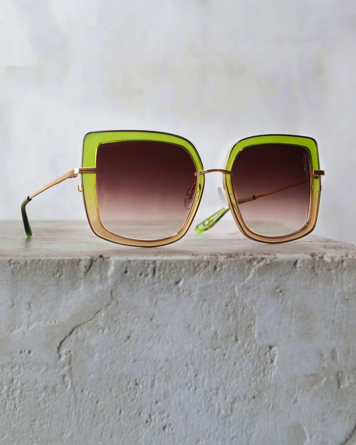 A pair of square-shaped sunglasses with gradient tinted lenses, a green frame on top, and gold accents, resting on a textured concrete surface.