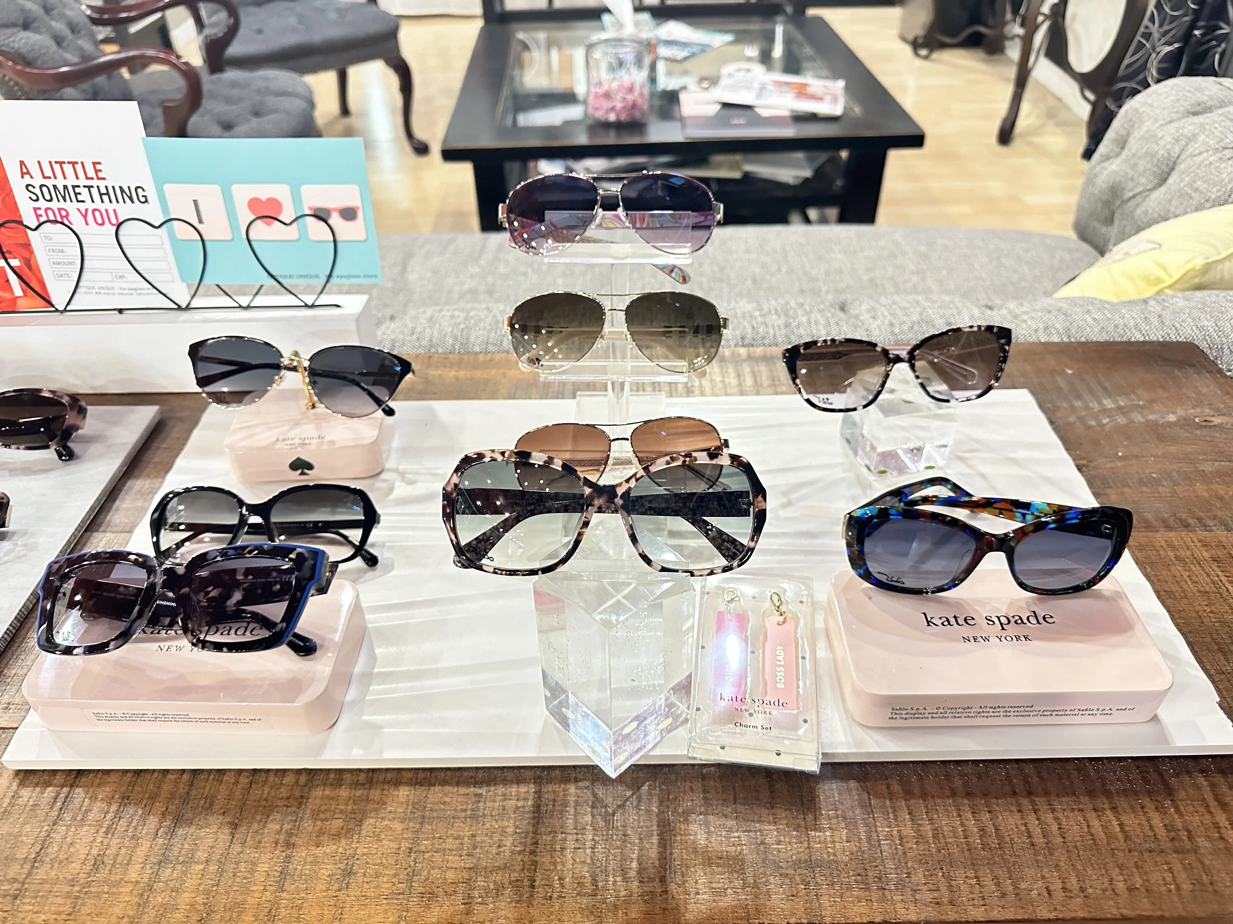Display of various sunglasses on a retail table at a store, with a white tray holding them, some on pink platform bases, and a small clear acrylic stand in the center. In the background, a gray couch with a yellow pillow and a black coffee table with magazines are visible.