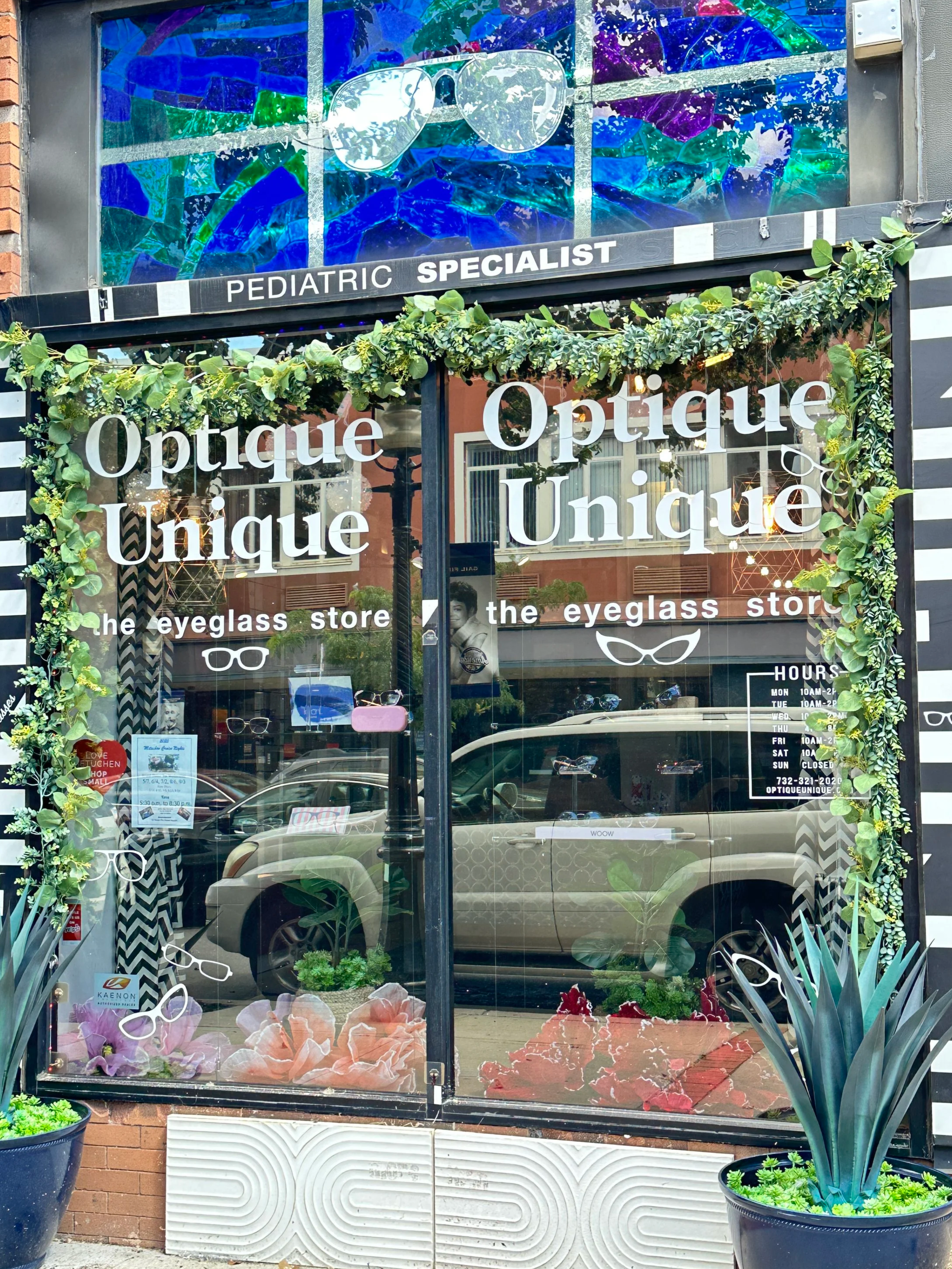 The storefront of Optique Unique, an eyeglass store, with large glass windows decorated with white text and sunglasses graphics, surrounded by plants. Reflected in the glass are parked cars and buildings across the street. Above the store, a sign reads 'Pediatric Specialist' with a colorful stained glass window featuring sunglasses.