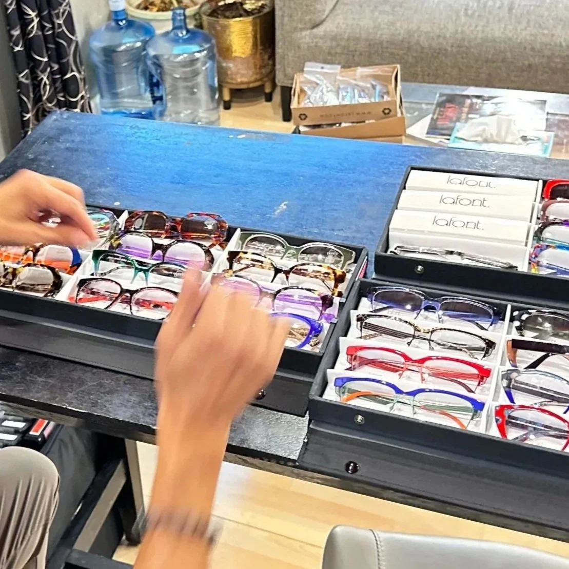 A person shopping for eyeglasses at a store, selecting from various sunglasses and optical frames displayed in open boxes on a table.