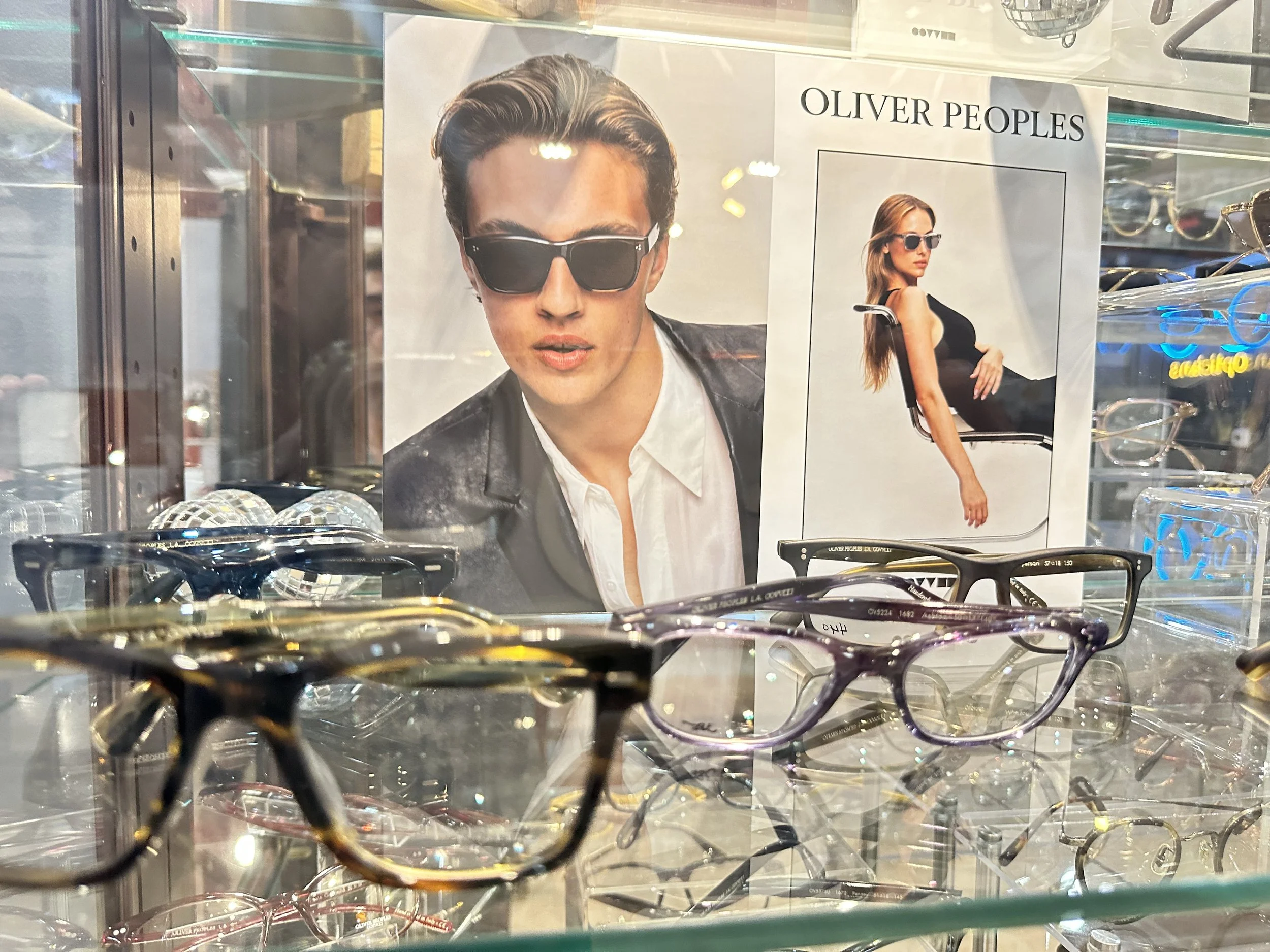 Display case with various eyeglasses and a poster promoting Oliver Peoples sunglasses featuring a man with dark sunglasses and a woman with dark sunglasses sitting in a chair.