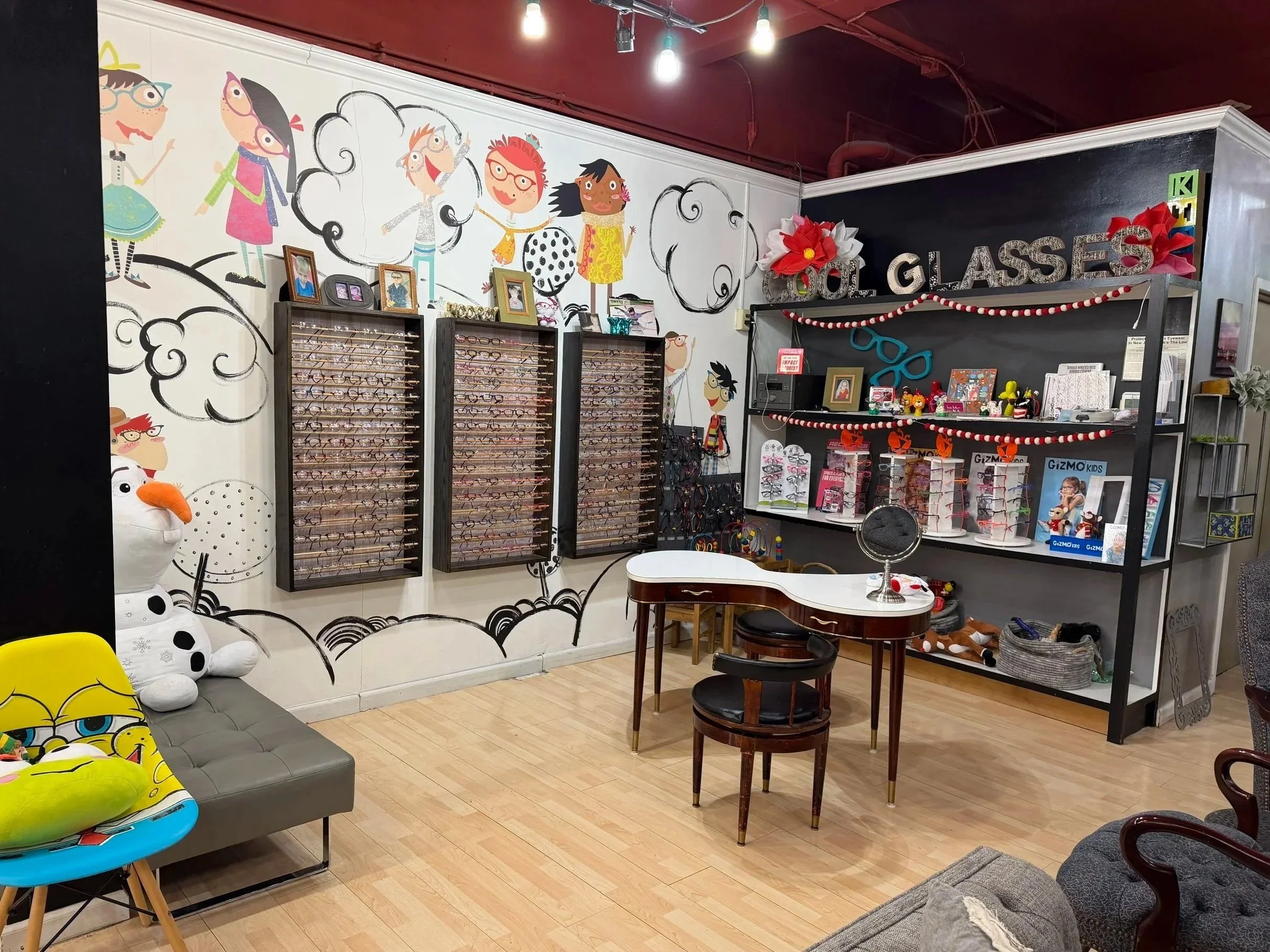 Optical store with wall mural of cartoon children, display cases of eyeglasses, a small table with a mirror, and shelves with eyeglass frames and accessories.