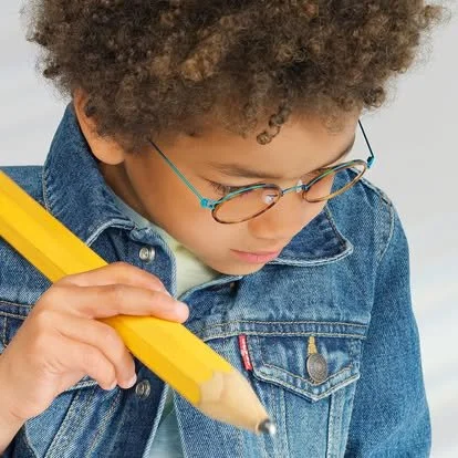A young boy with curly hair wearing glasses and a denim jacket, holding a large yellow pencil and looking down at what he's writing.