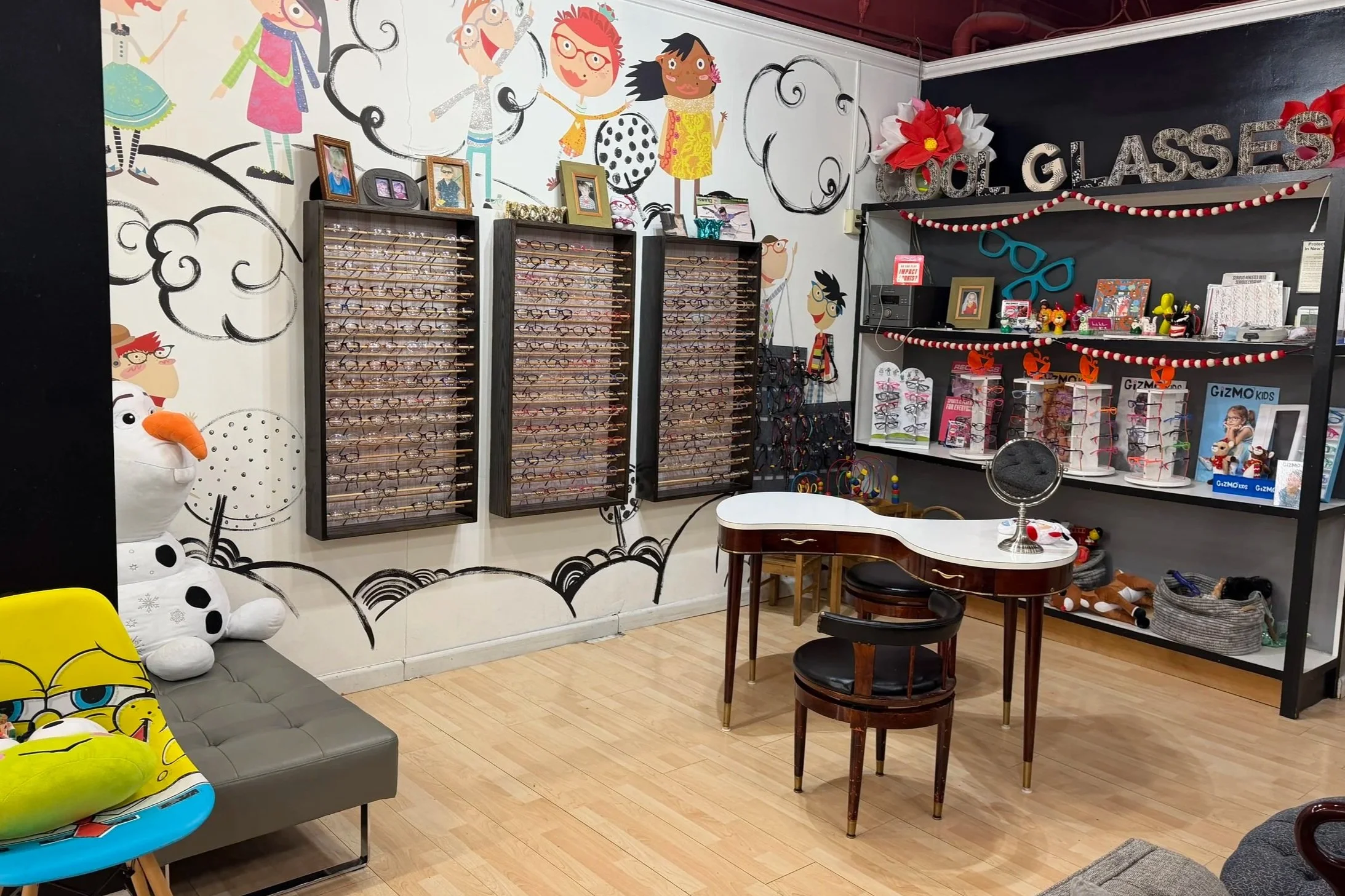 Eyewear store with glasses display cases, a small white and brown table with two chairs, wall art with cartoon characters, shelves with toys and accessories, and decorative items.