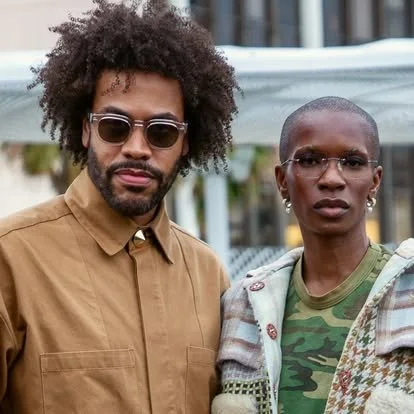 A man with curly hair and sunglasses and a woman with short hair and glasses standing together outdoors.