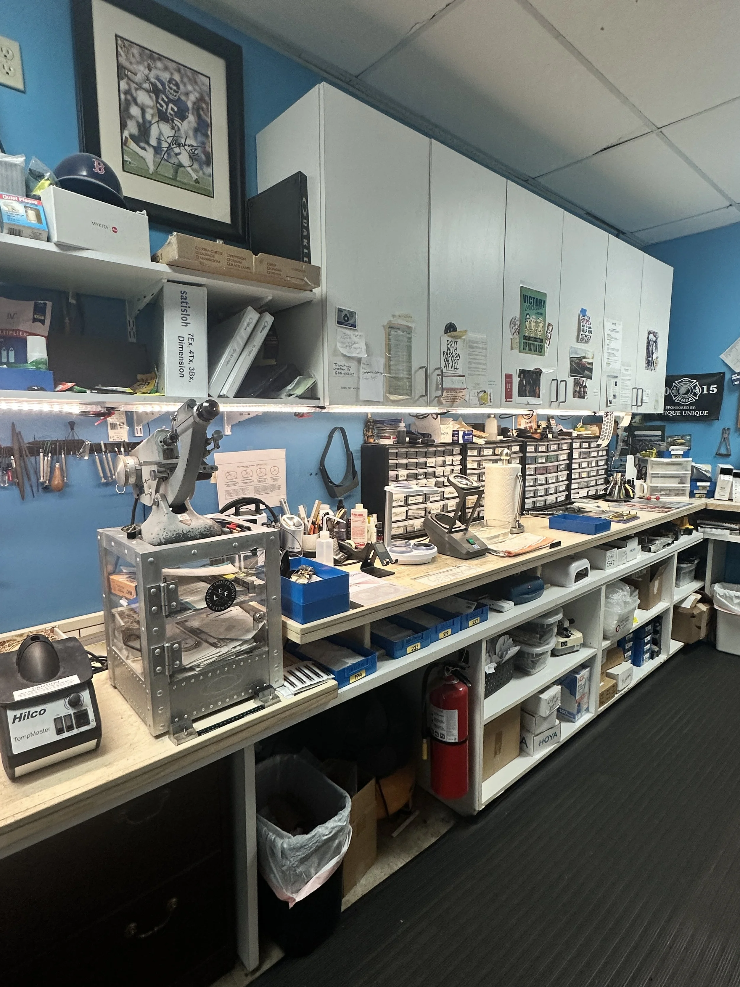 Workshops tools and supplies organized on a workbench in a workshop or repair shop. Includes a microscope, a fire extinguisher, drawers with small parts, and various tools on the wall.