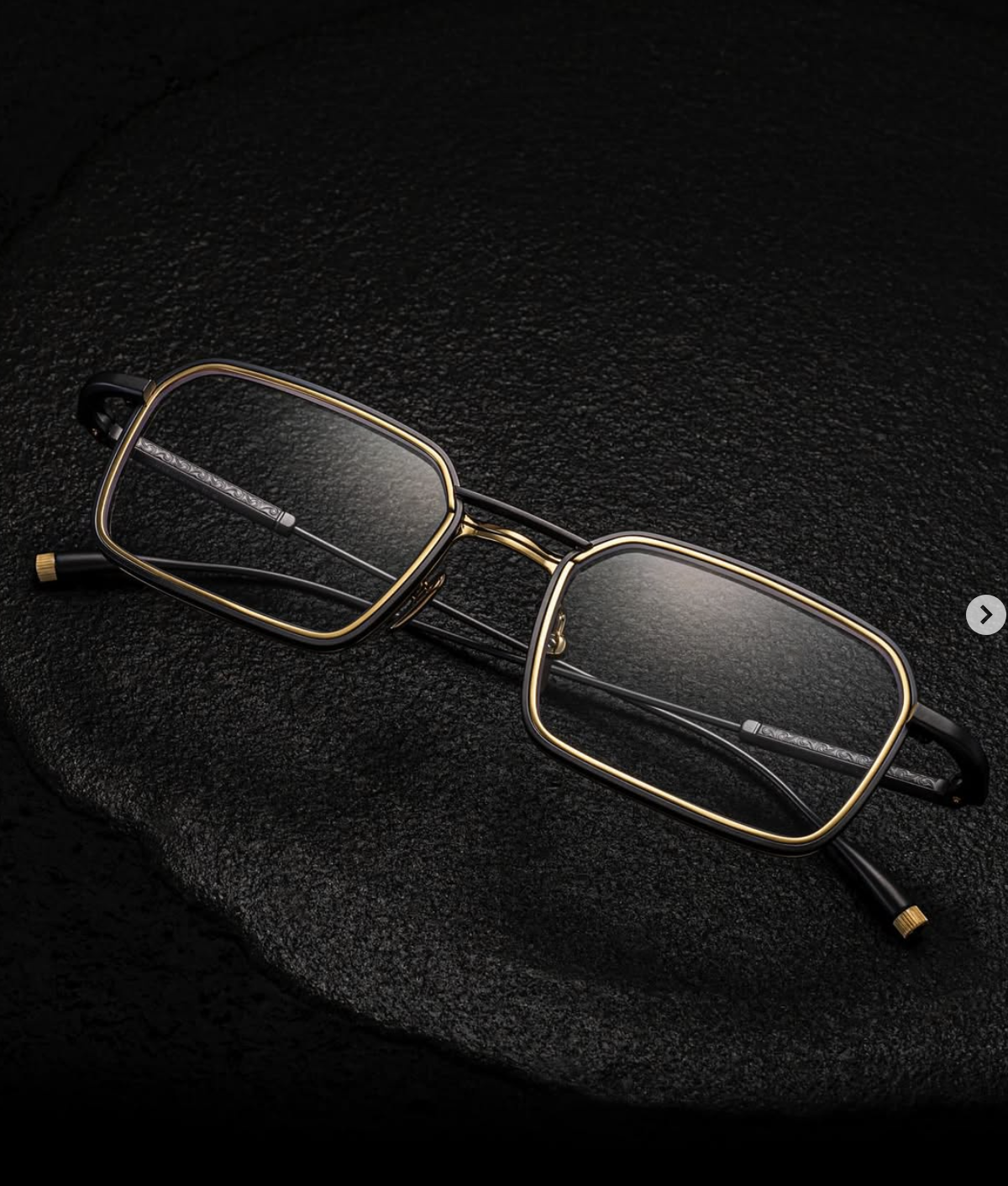 A pair of rectangular eyeglasses with thin black and gold frames resting on a black textured surface.