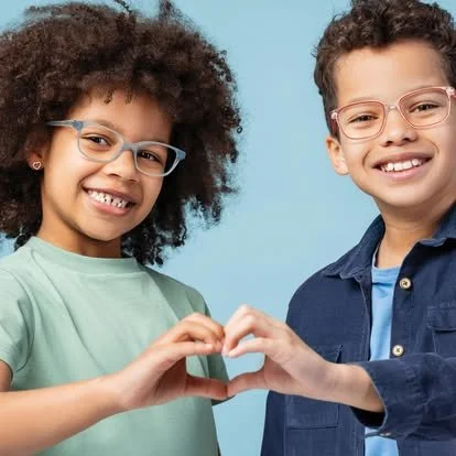 Two children, a girl with curly hair and a boy, stand side by side against a blue background, smiling and forming a heart shape with their hands.