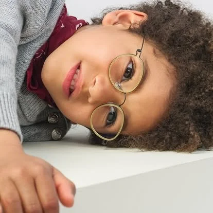 Child with curly hair and glasses lying on a white surface, looking at the camera.
