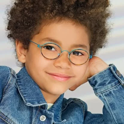 Child with curly hair wearing glasses and a denim jacket, smiling outdoors.
