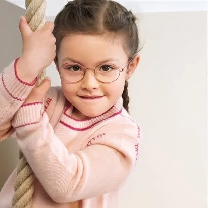 A young girl with glasses and braided hair, wearing a pink sweater, holding onto a thick rope with both hands, standing indoors.