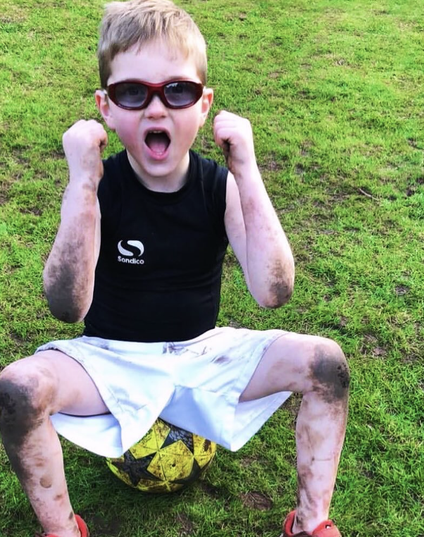 A young boy sitting on a yellow and black soccer ball on a grassy field, wearing sunglasses, a black sleeveless shirt, white shorts, and red shoes. He has dirt or mud smudges on his arms, legs, and face, and he is flexing his arms with an excited exp