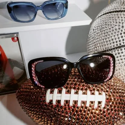 Pair of black sunglasses with pink rhinestones on the frame, resting on a brown textured clutch or purse, with a white decorative item and blue sunglasses in the background.