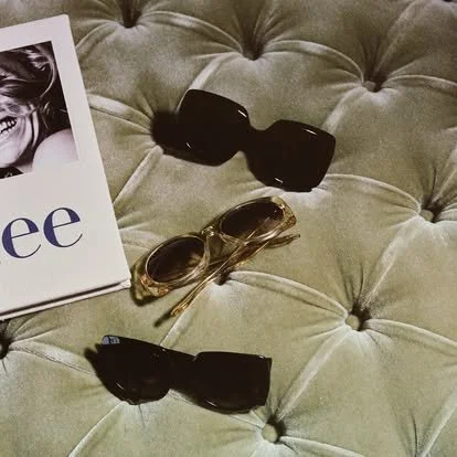 Two pairs of sunglasses on a tufted, beige fabric surface, with part of a magazine or flyer showing a smiling woman and some text.