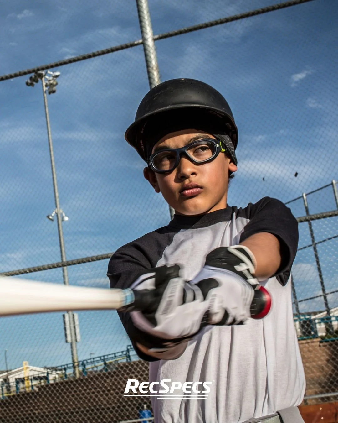 Young boy wearing helmet and goggles holding a baseball bat at a baseball field.