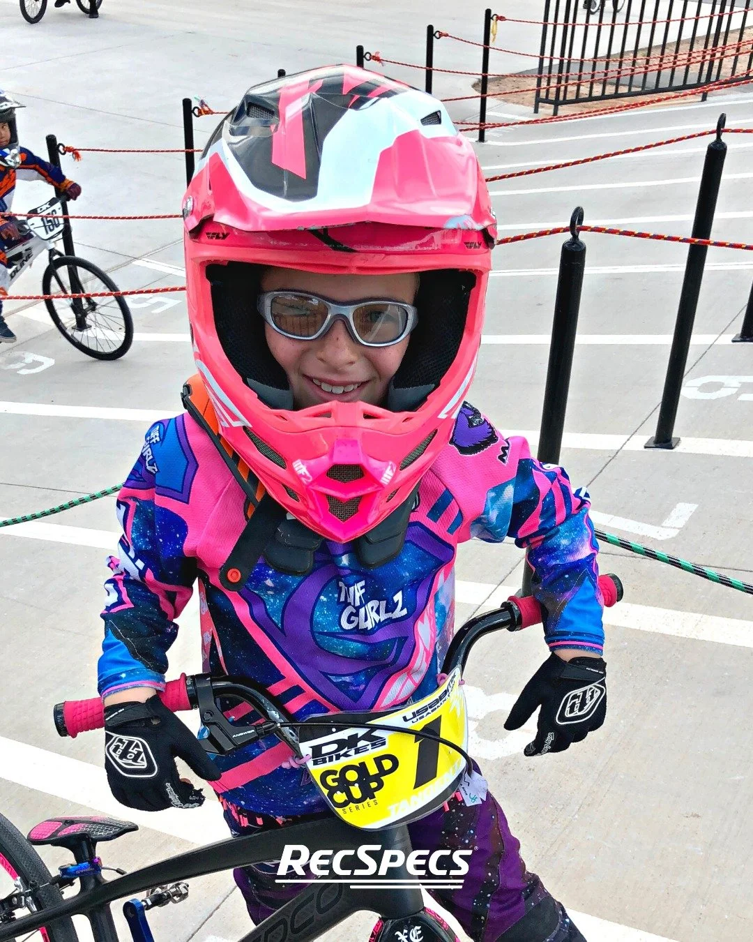 A young girl wearing a pink helmet and goggles, dressed in colorful motocross gear, sitting on a bicycle in a parking lot, smiling at the camera.