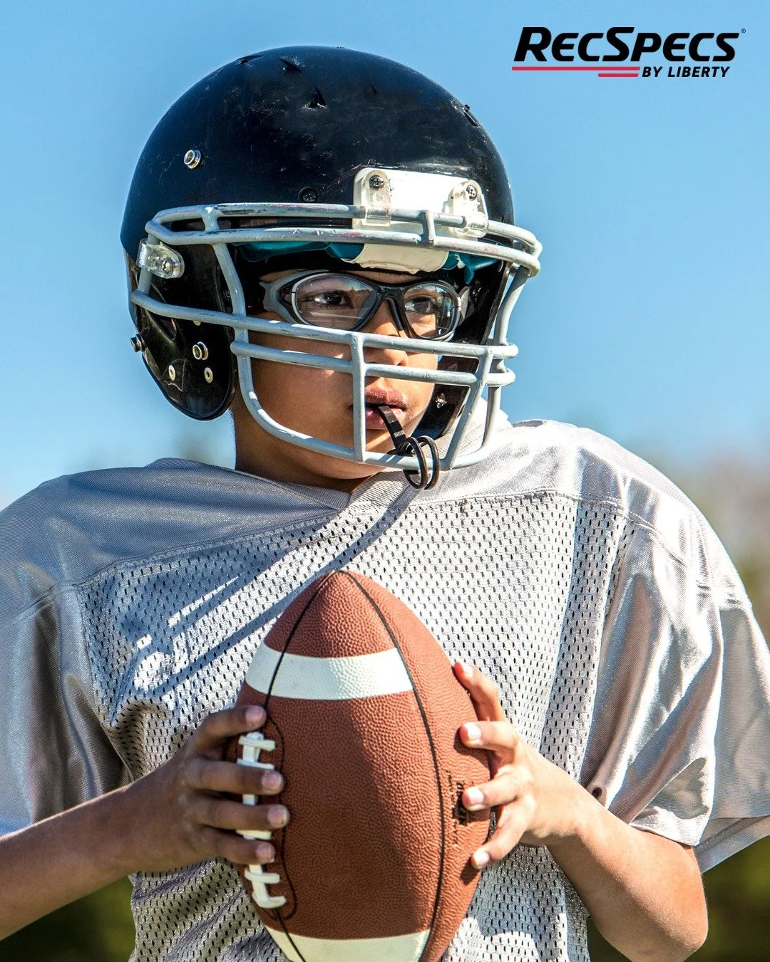 A young football player wearing a black helmet, glasses, and a white jersey holding a brown football with white stripes outdoors.