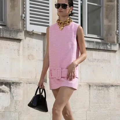 A woman in a pink sleeveless dress with a waist belt, wearing sunglasses and a gold chain necklace, holding a black handbag, standing outside near a building with window shutters.
