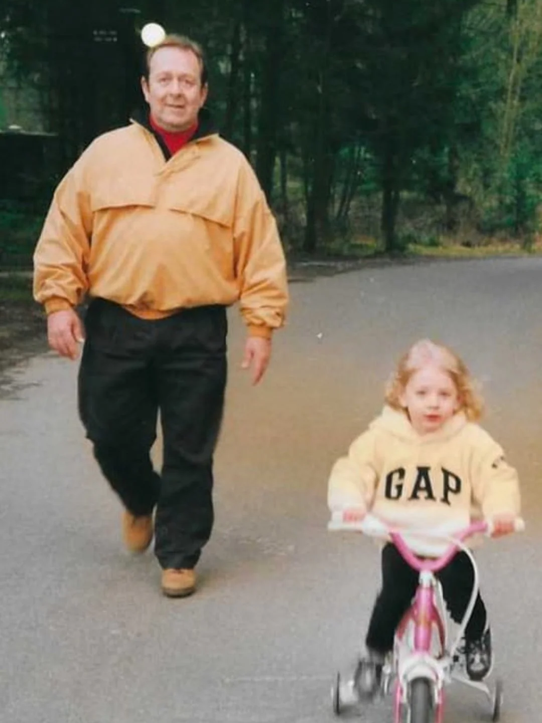 A man and a young girl are walking outdoors in a wooded area. The man is wearing a windbreaker, black pants, and tan shoes. The girl is riding a small pink bicycle with training wheels and is wearing a cream-colored hoodie with the brand name 'GAP' on it, black pants, and sneakers.