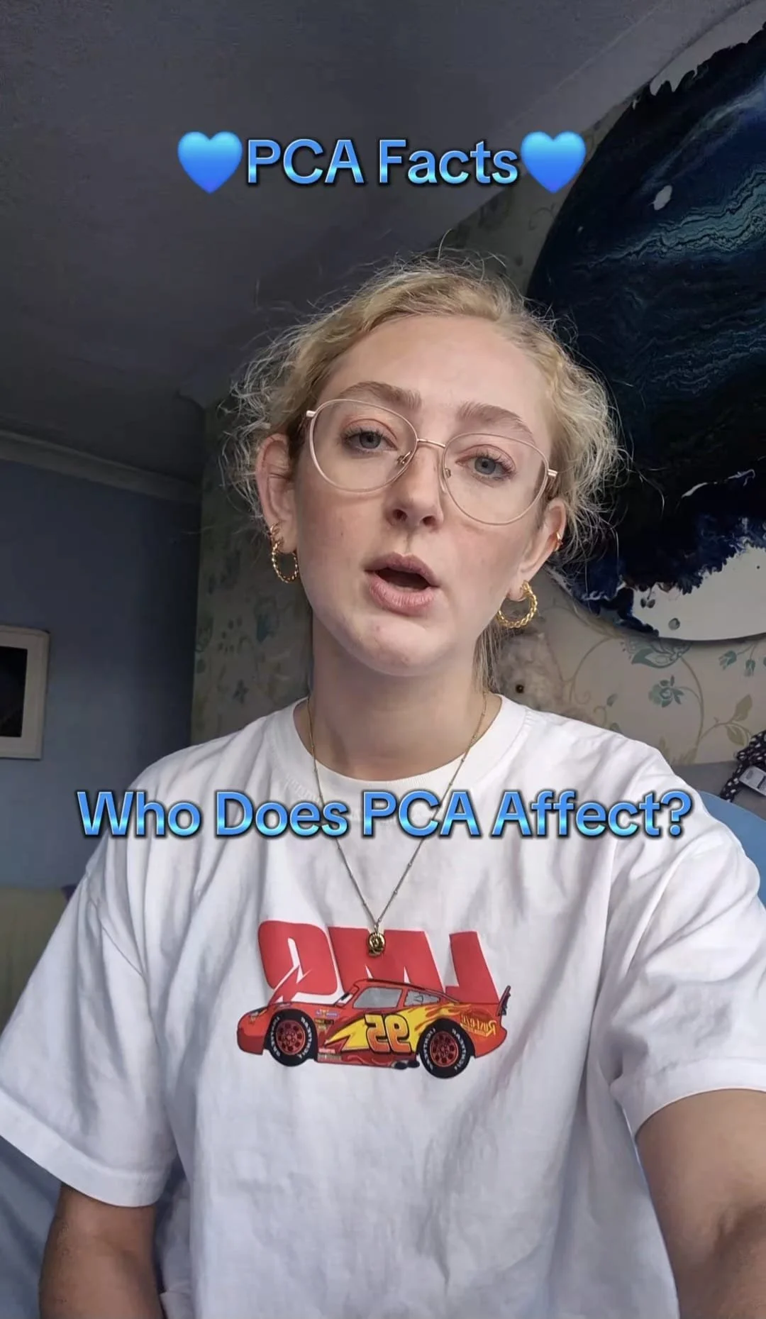 A woman with short blonde hair, glasses, and earrings takes a selfie in a room with a large abstract painting on the wall behind her, wearing a white T-shirt with a red and yellow Lightning McQueen car graphic and a caption asking about PCA effects.