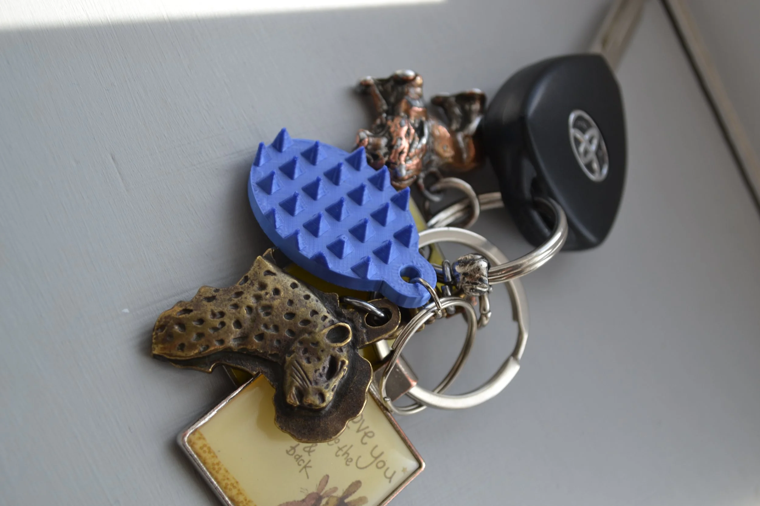 A collection of keychains including a blue textured dinosaur, a metal dog, and other characters, hanging on a keyring with an attached decorative card and a black car key.