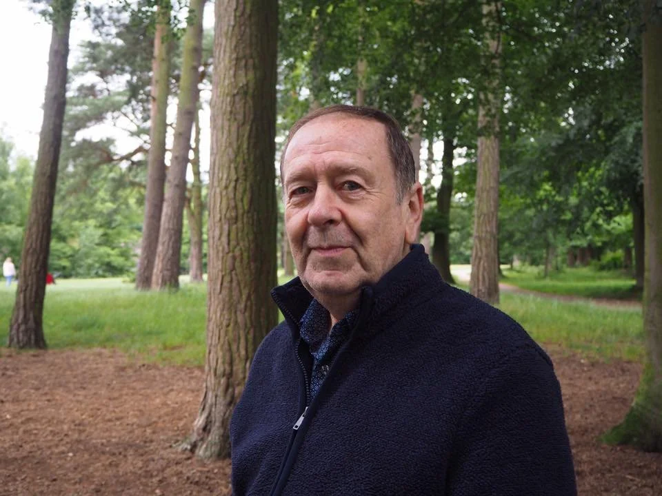 A middle-aged man with dementia wearing a navy fleece jacket standing in a wooded park.