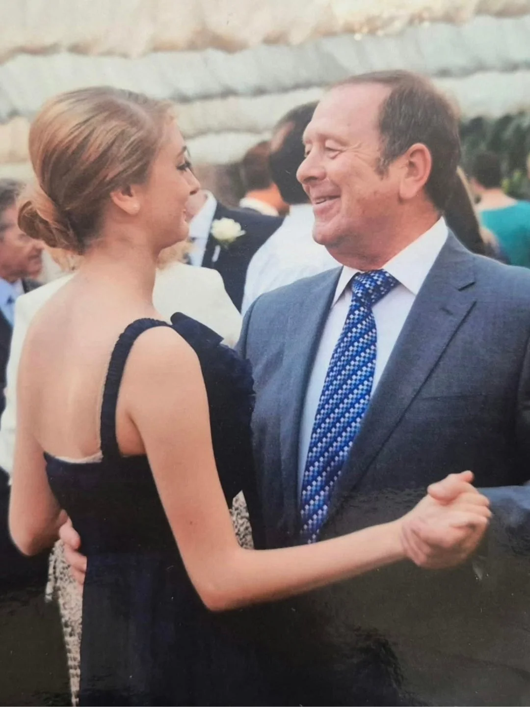 A man and a woman dancing at an event. Both are smiling. The man wears a nice suit with a blue tie and the young lady has an asymmetrical dress and her hair is up.