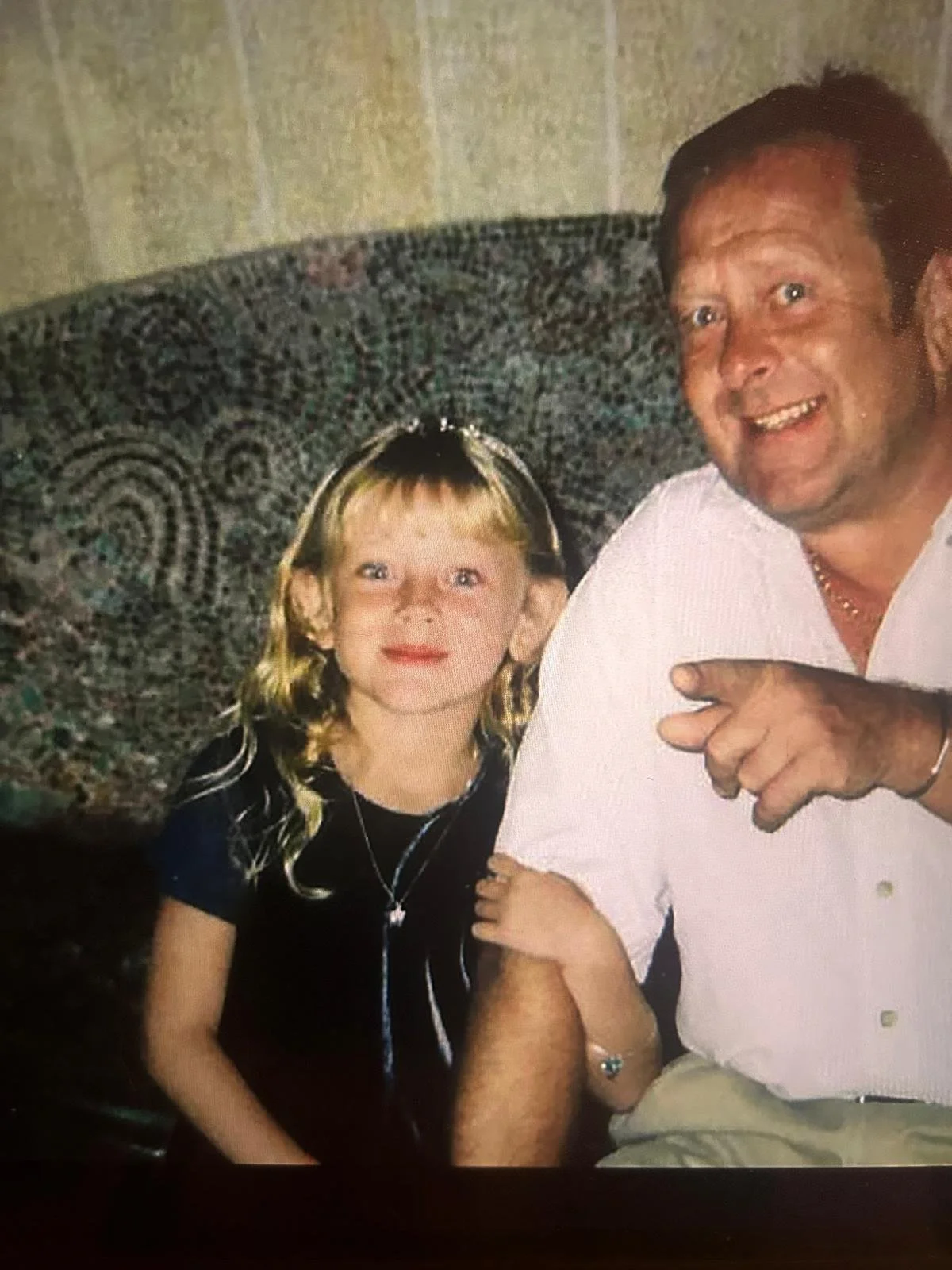 A man and a young girl are sitting on a patterned sofa, smiling at the camera. The girl has long blonde hair, and the man has dark hair and a beard. The man is pointing towards the girl.