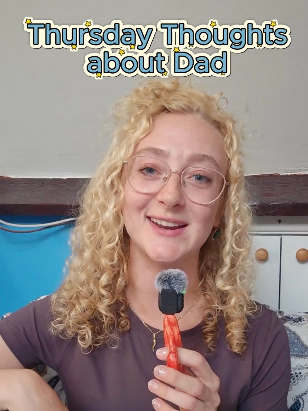 A woman with curly blonde hair, glasses, and a dark shirt holding a microphone with a furry windscreen. Text overlay reads 'Thursday Thoughts about Dad' with small star accents.