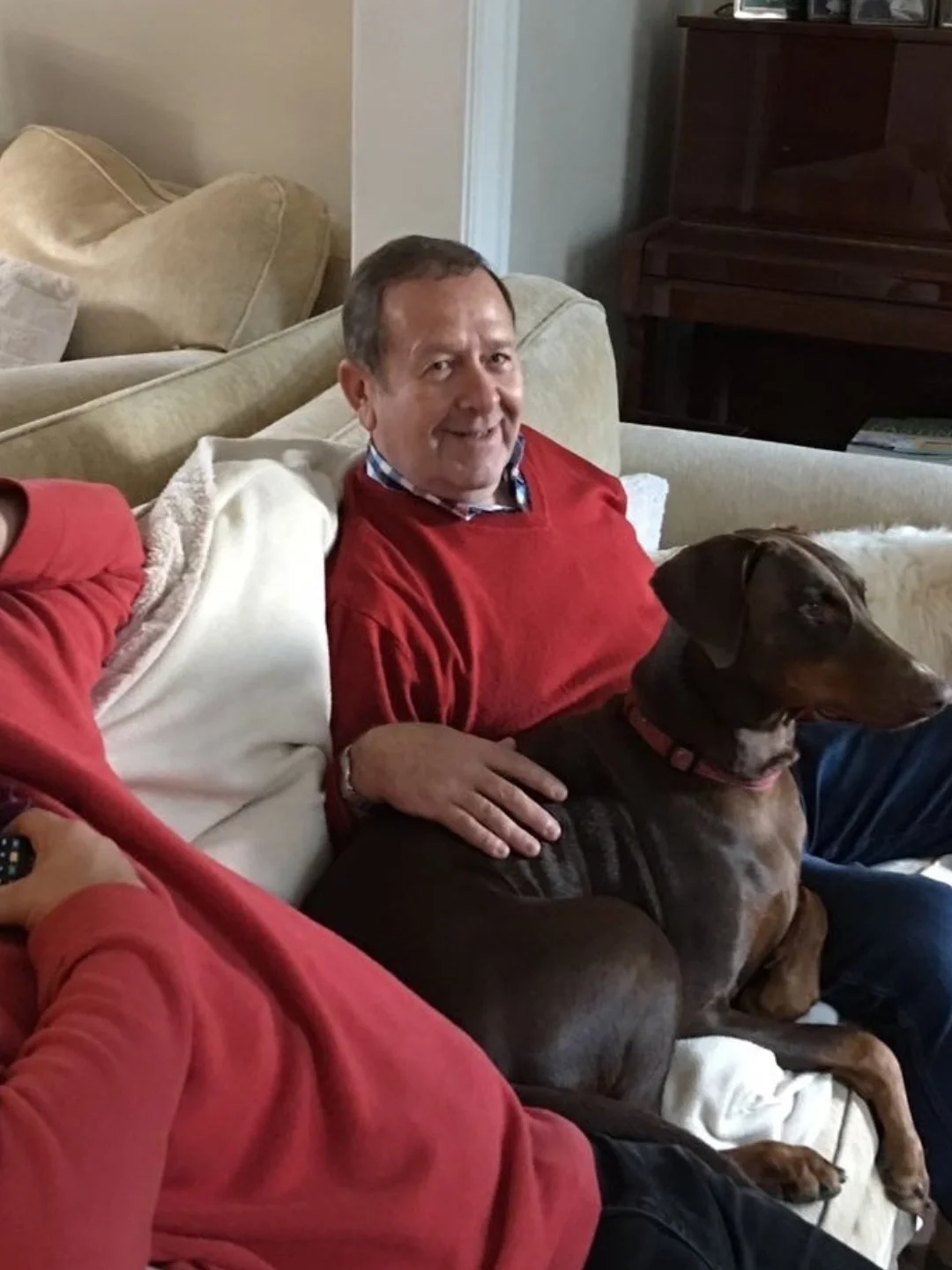 A man in a red sweater sitting on a couch with a brown dog on his lap, smiling at the camera. Another person in a red shirt is partially visible, holding a remote.