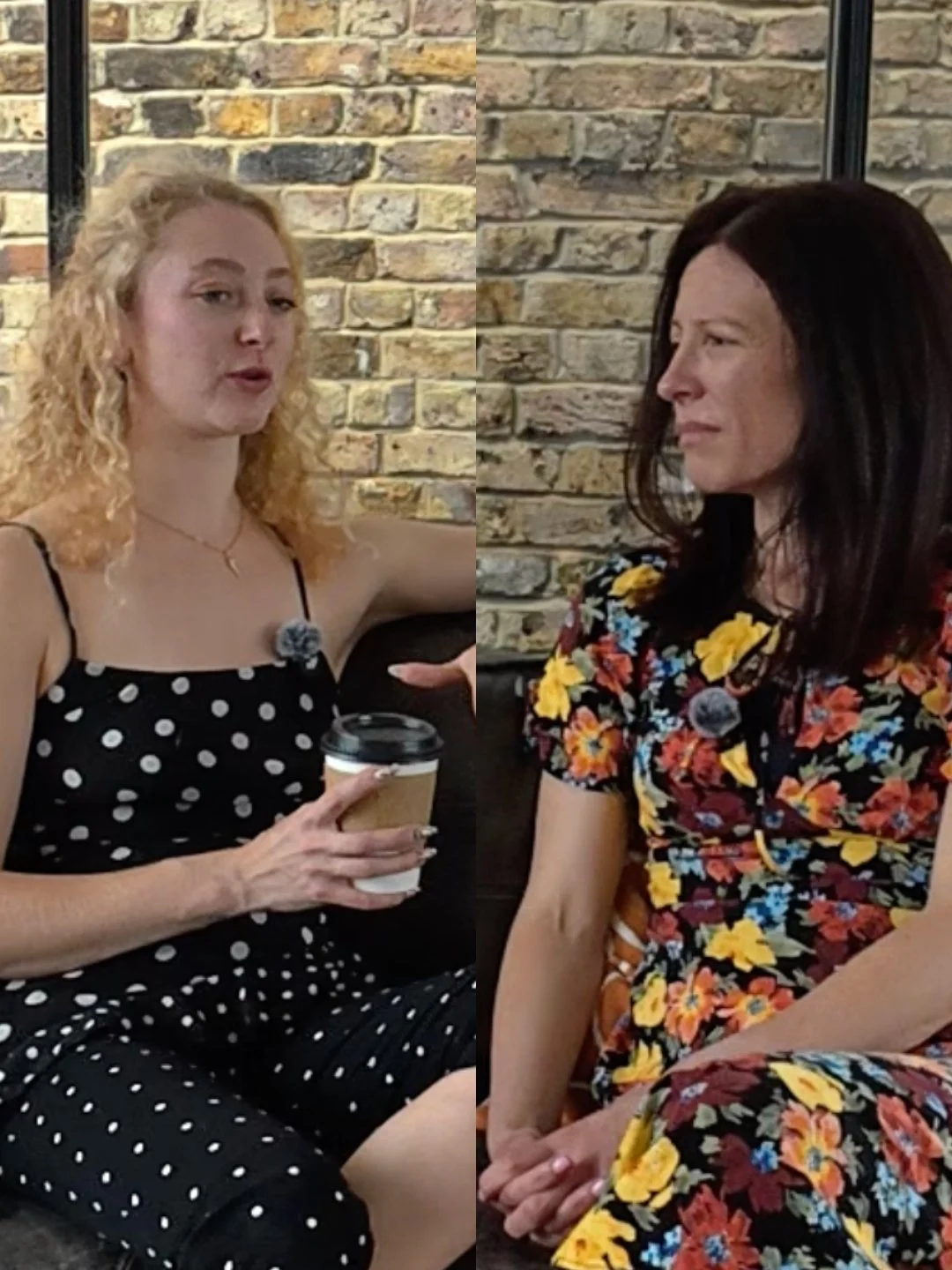 Two women are sitting in front of a brick wall, engaged in conversation. The woman on the left has curly blonde hair and is wearing a black dress with white polka dots, holding a cup. The woman on the right has shoulder-length dark hair and is wearing a colorful floral dress.