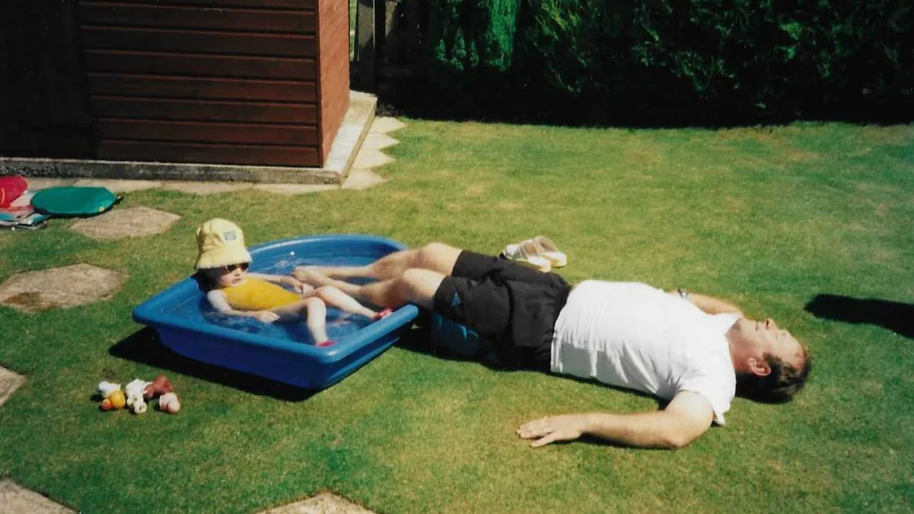 A man lying on green grass next to a small child in a plastic kiddie pool, both relaxing on a sunny day.