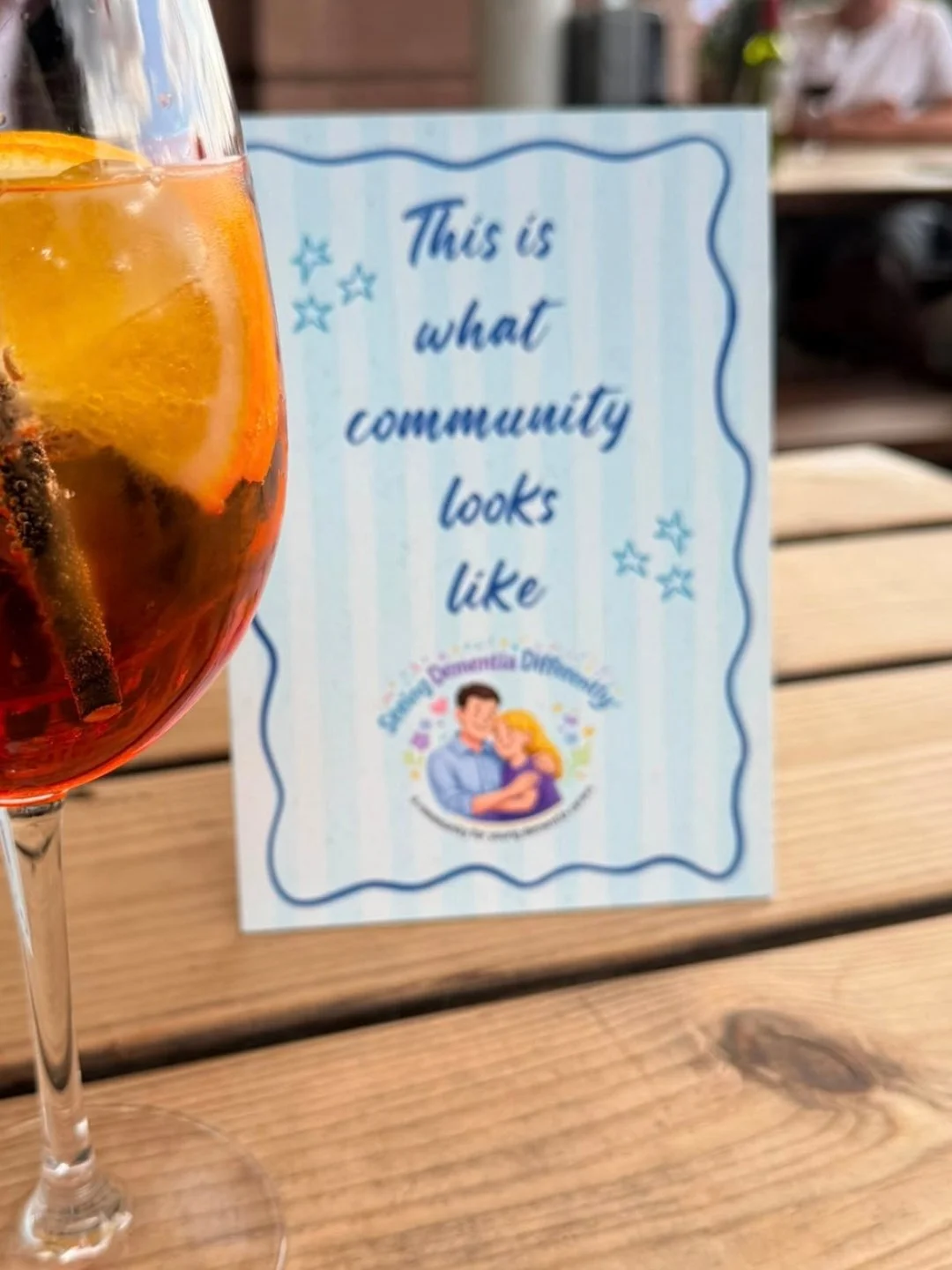 A fruit drink with an orange slice next to a sign that reads 'This is what community looks like'. Advertising a Seeing Dementia Differently young adult dementia carers meet up.