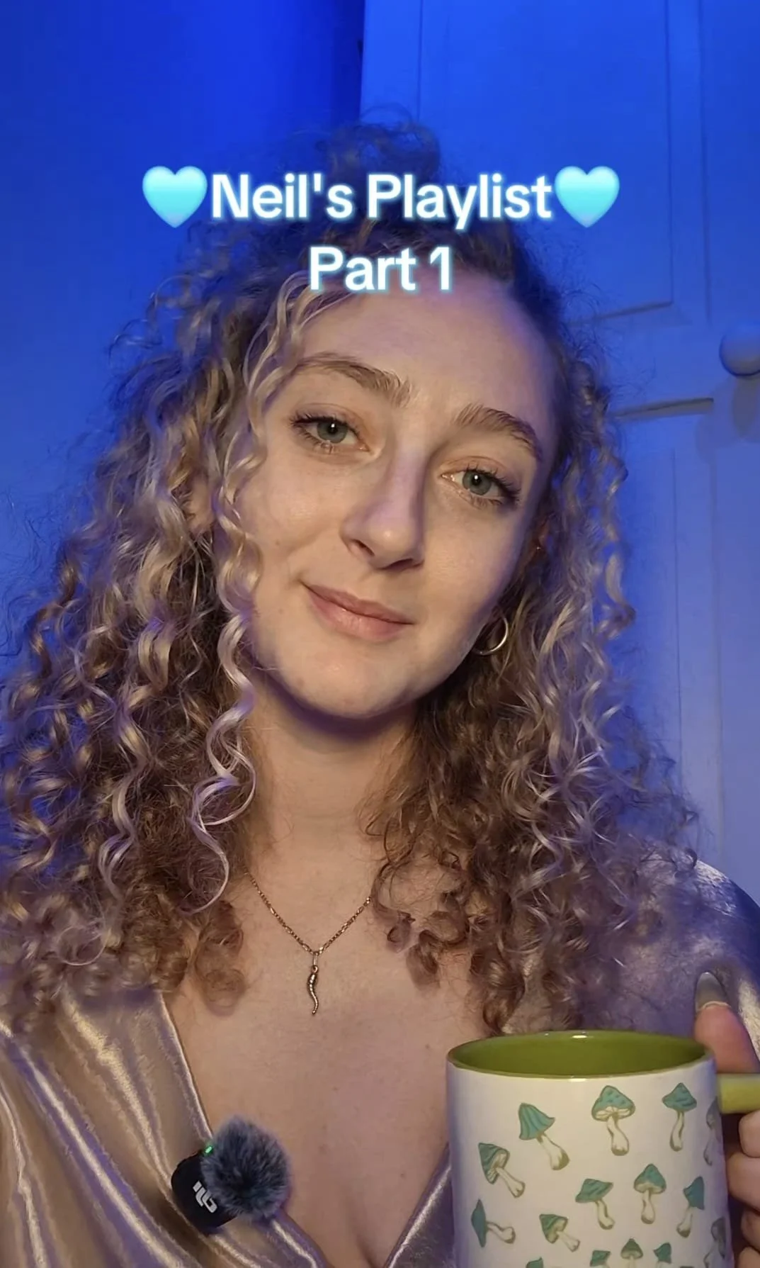 A young woman with curly blonde hair holding a mug with mushroom illustrations, smiling at the camera, with text overlay saying 'Neil's Playlist Part 1' and blue heart icons.