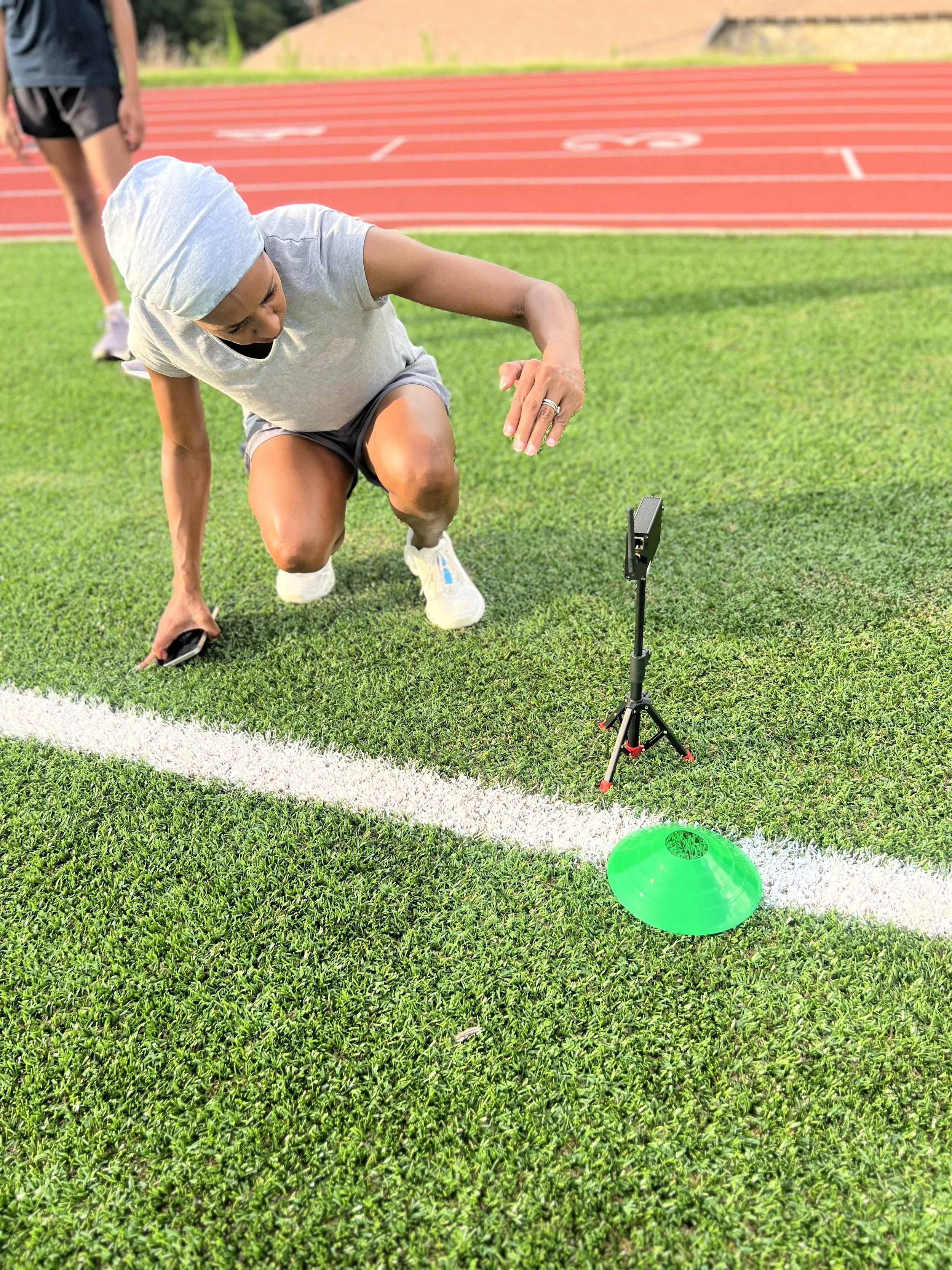 Athletic woman kneeling on a track field, adjusting a device near a green cone, with a person walking in the background and a red running track.