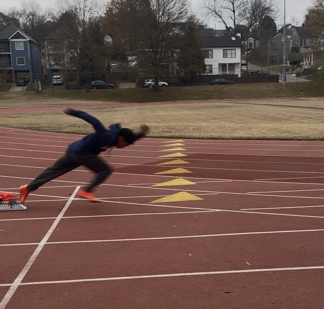 A person in dark clothing is starting a sprint on a red running track, leaning forward with one arm extended behind and the other arm bent in front, wearing bright orange shoes. The track has yellow starting blocks and numbered lanes. In the background, there are residential houses, leafless trees, and a cloudy sky indicating a late fall or winter day.