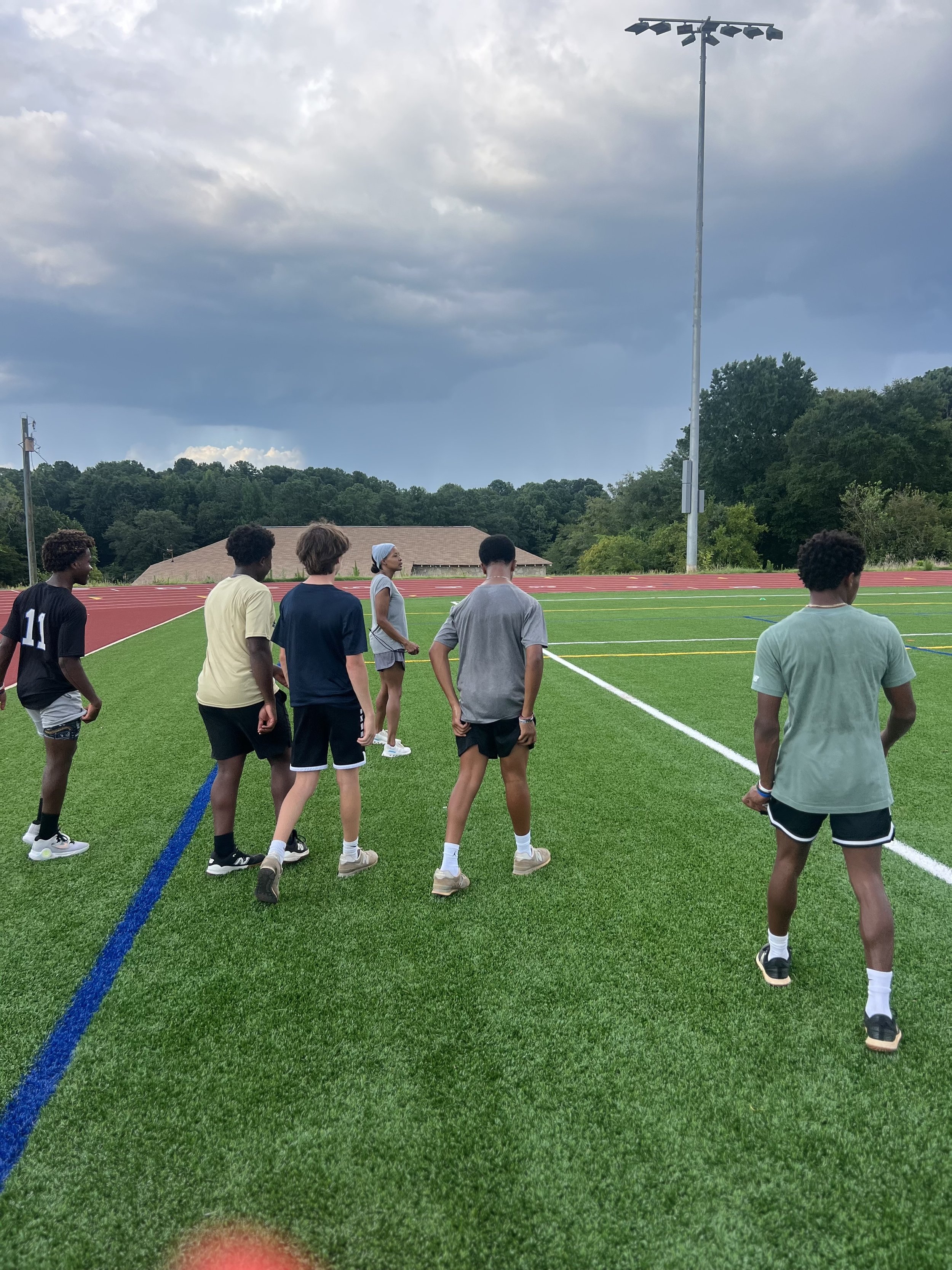 A group of young people standing on a green athletic field, facing away from the camera, preparing for a run or exercise, with trees, a building, and a cloudy sky in the background.