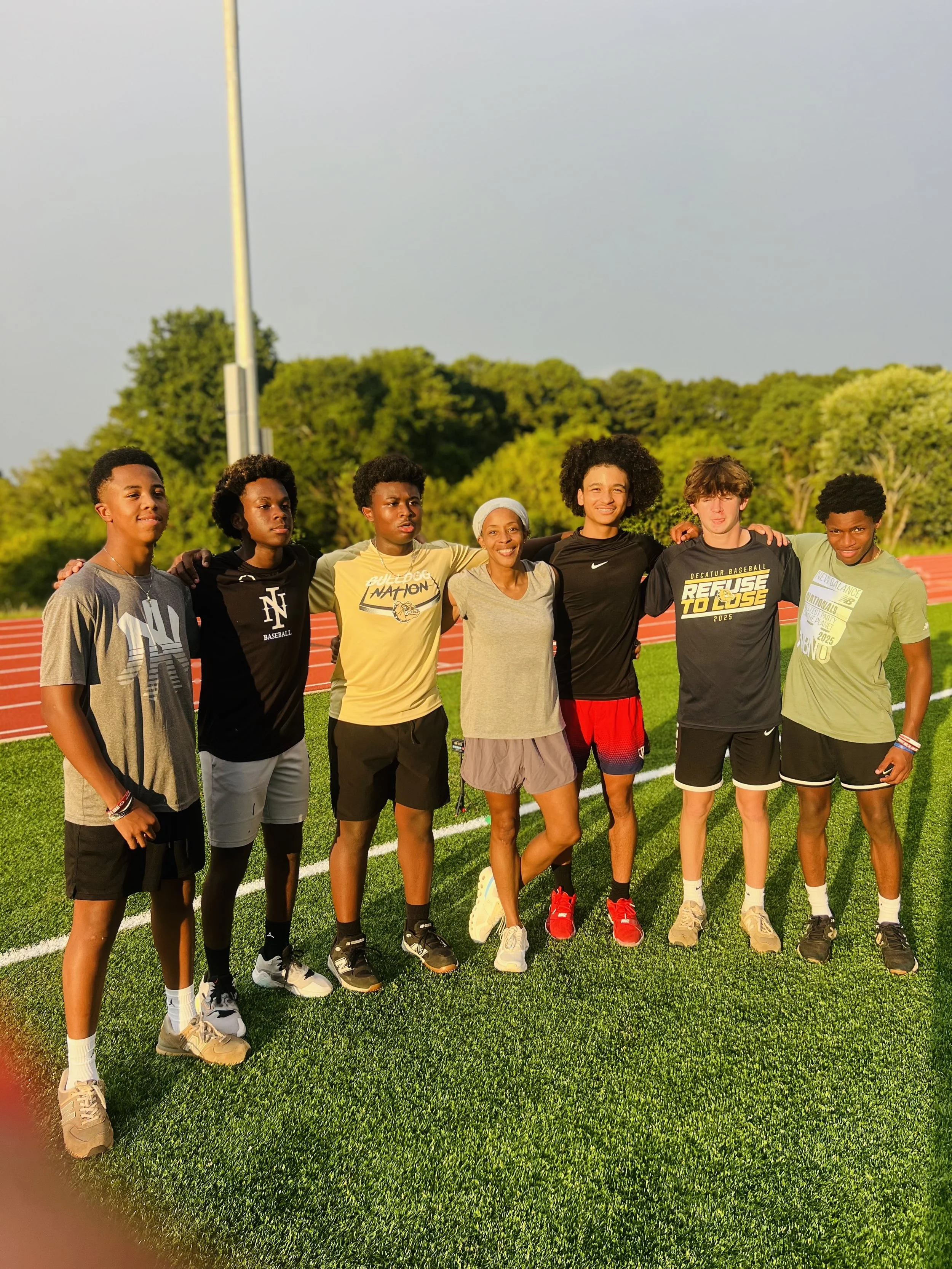 A group of seven young athletes standing under a bright sky on a green track field with trees in the background, smiling and posing for a photo.