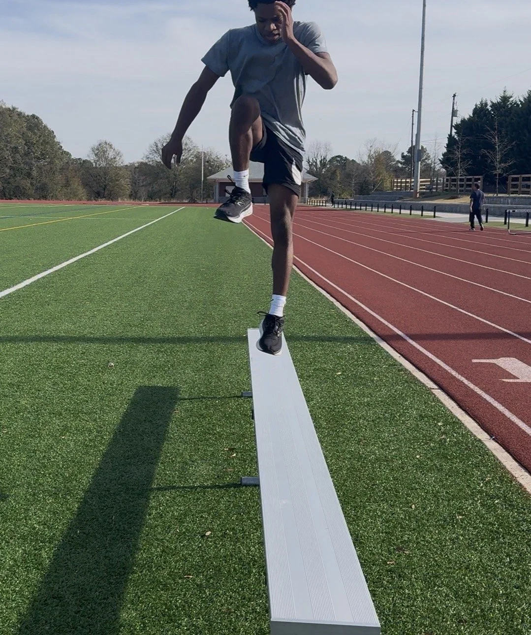 Young man balancing on a narrow white beam on a track field, stretching his leg and holding his head, with a running track and some people in the background.