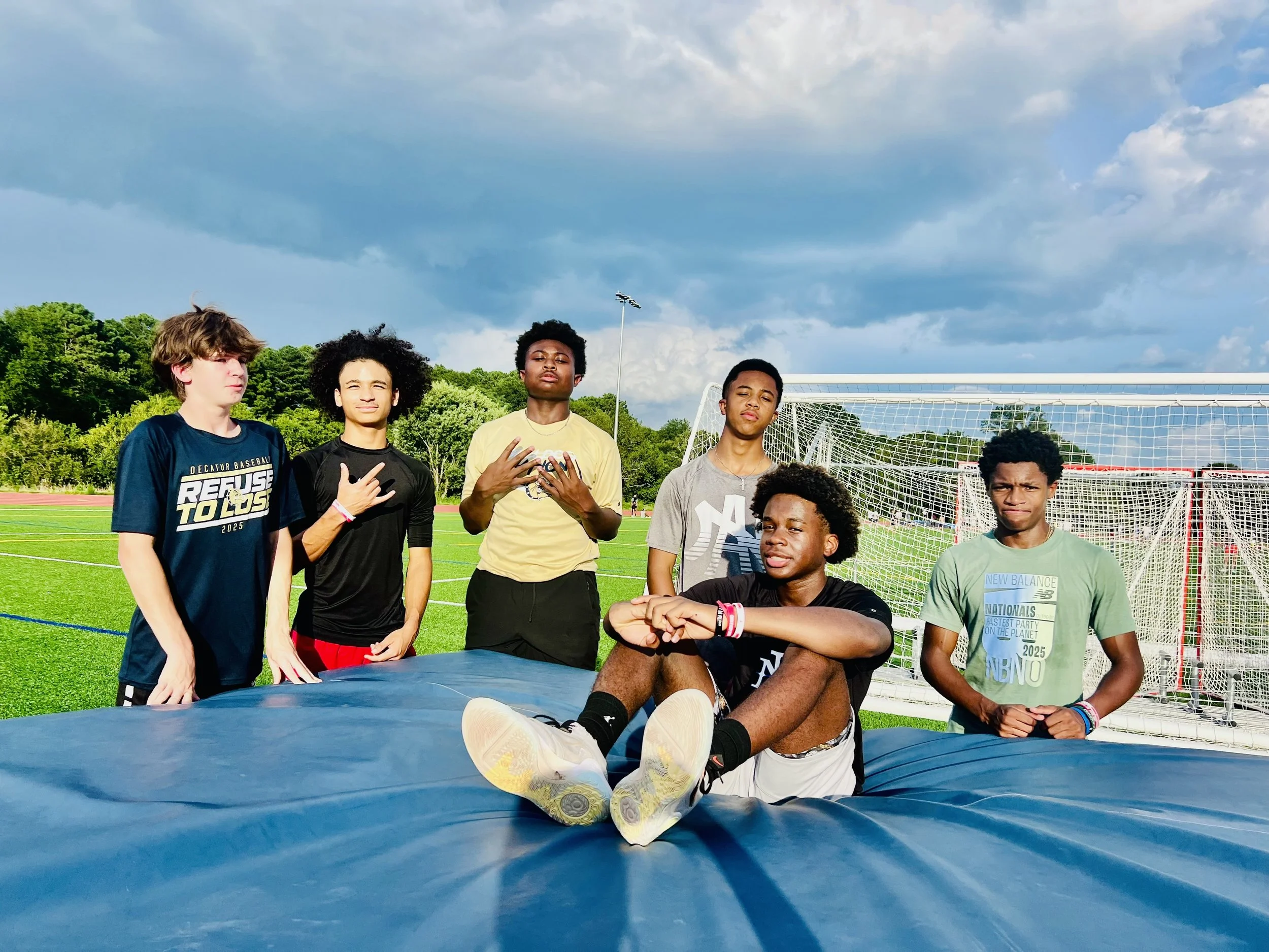 Group of six teenagers on a sports field, with one boy sitting on a large tarp and the others standing behind him, during daytime with cloudy sky.