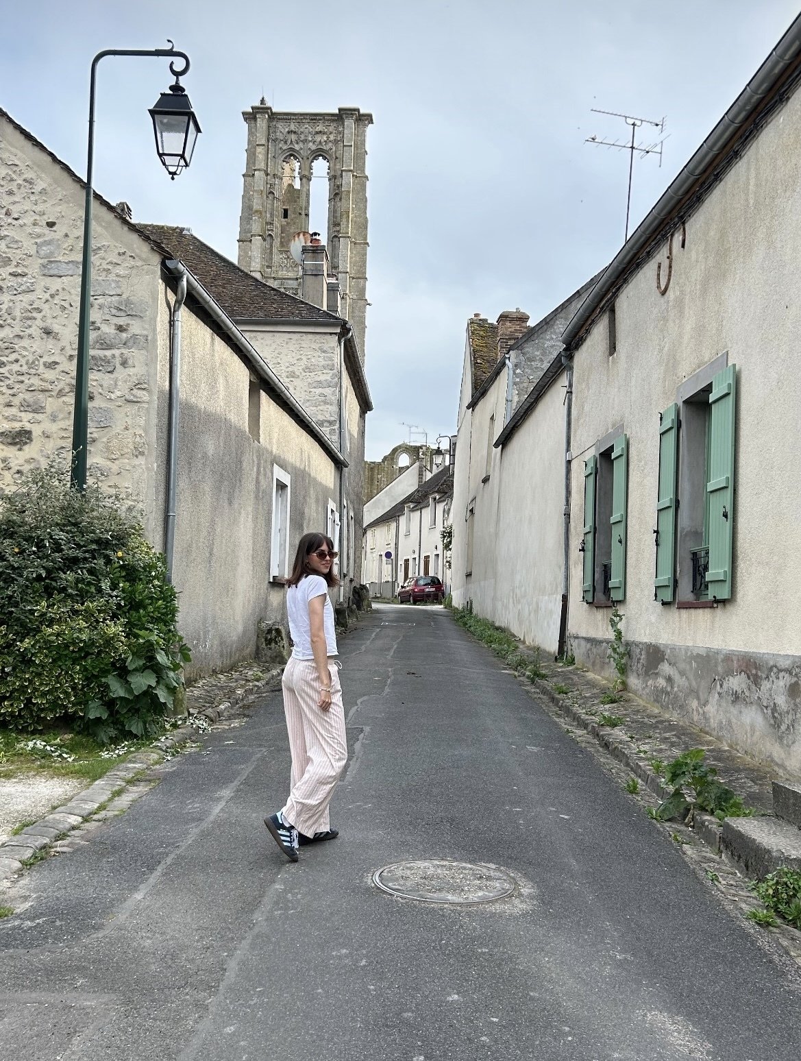 A woman in sunglasses wearing a white t-shirt and striped pink pants walking on a narrow French street with old stone buildings and green shutters, with a church bell tower in the background.