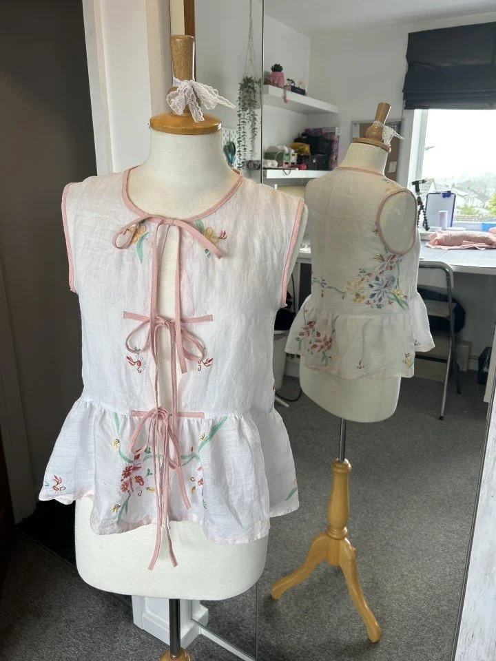 A white sleeveless women's top with pink embroidery and tie closures on a mannequin, reflected in a mirror in a sewing or craft room.