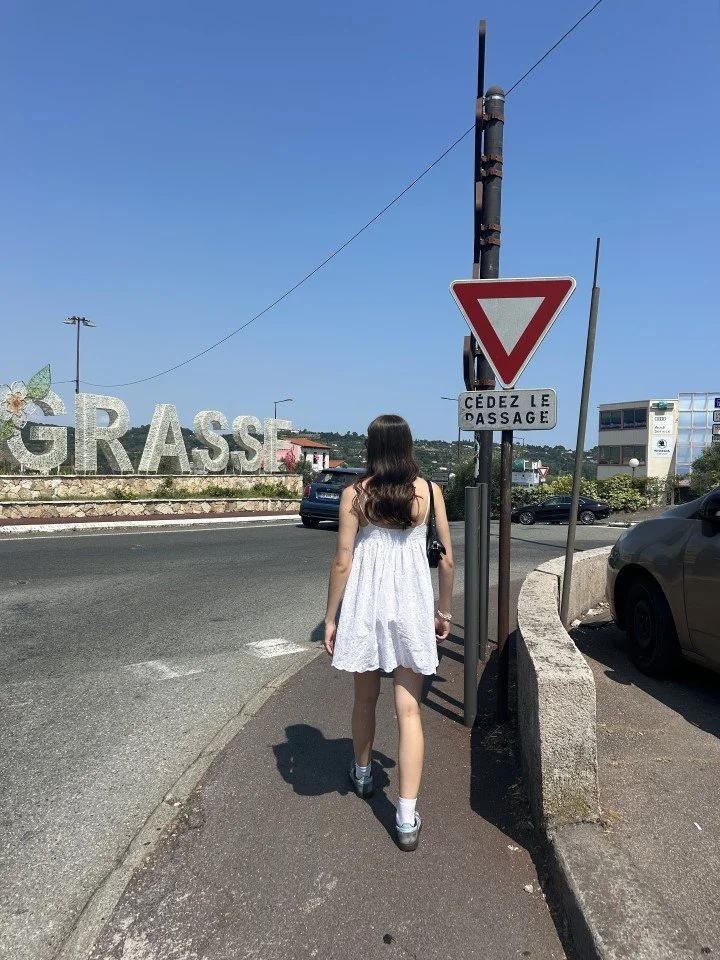A young woman in a white dress walking on the sidewalk next to a street, with a traffic yield sign and a sign reading "Cédez le passage." In the background, there's a large sign spelling "GRASSE" with a flower decoration, and some buildings and parked cars under a clear blue sky.
